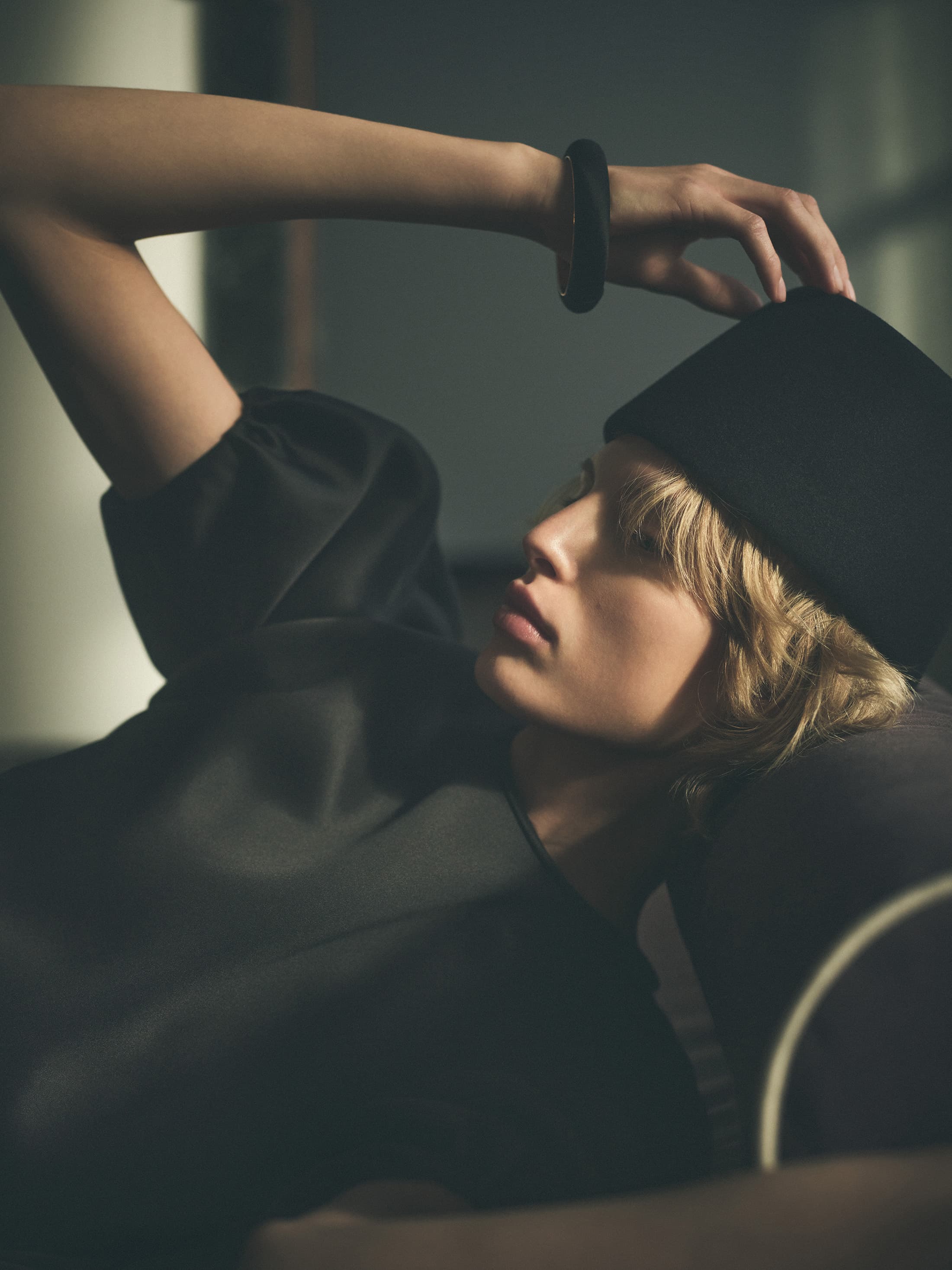 Model in a black pillbox hat and dark top reclining indoors with one hand raised to her head.