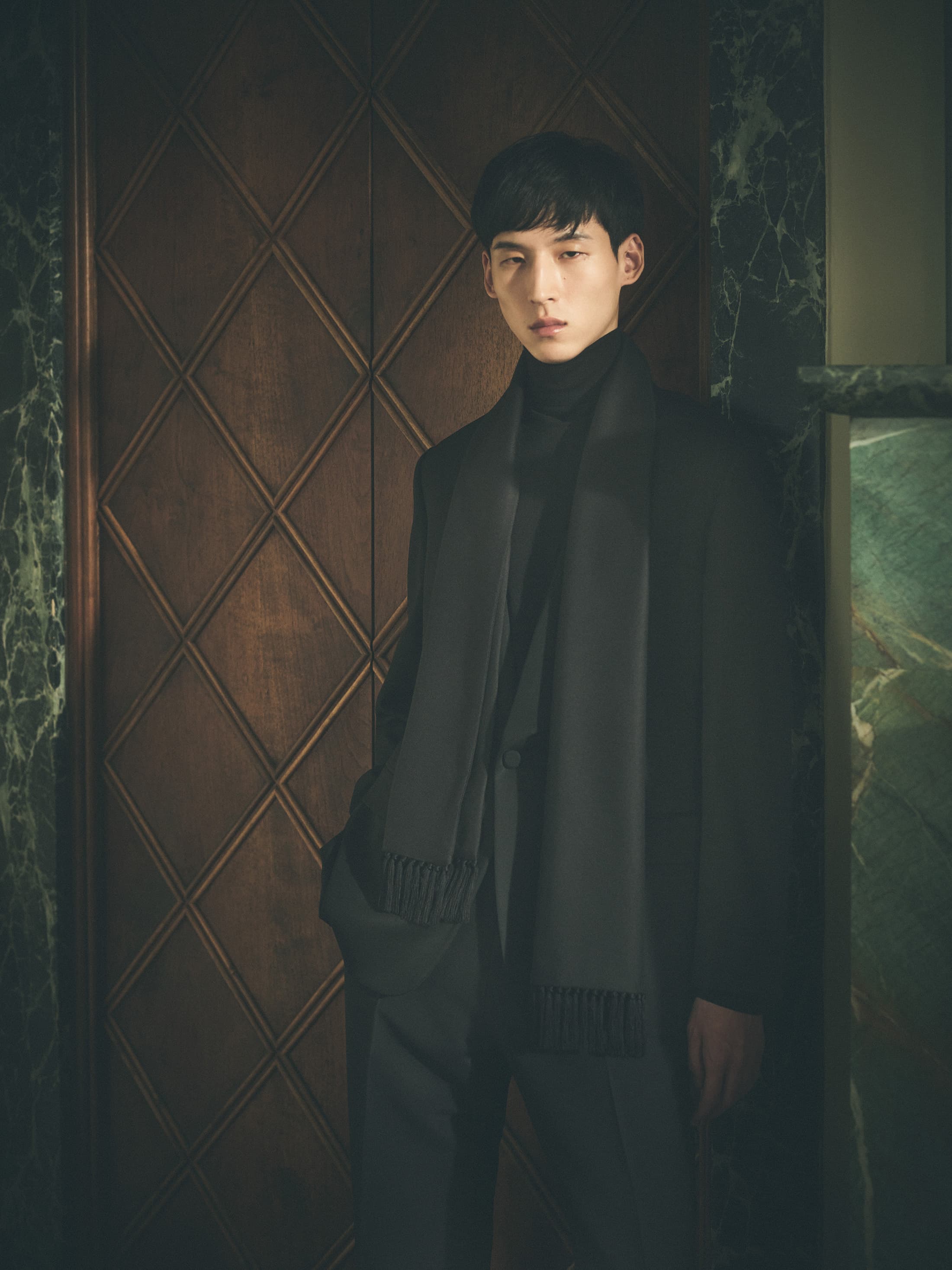 Man wearing a dark suit, turtleneck, and scarf standing in front of a wood-paneled door.