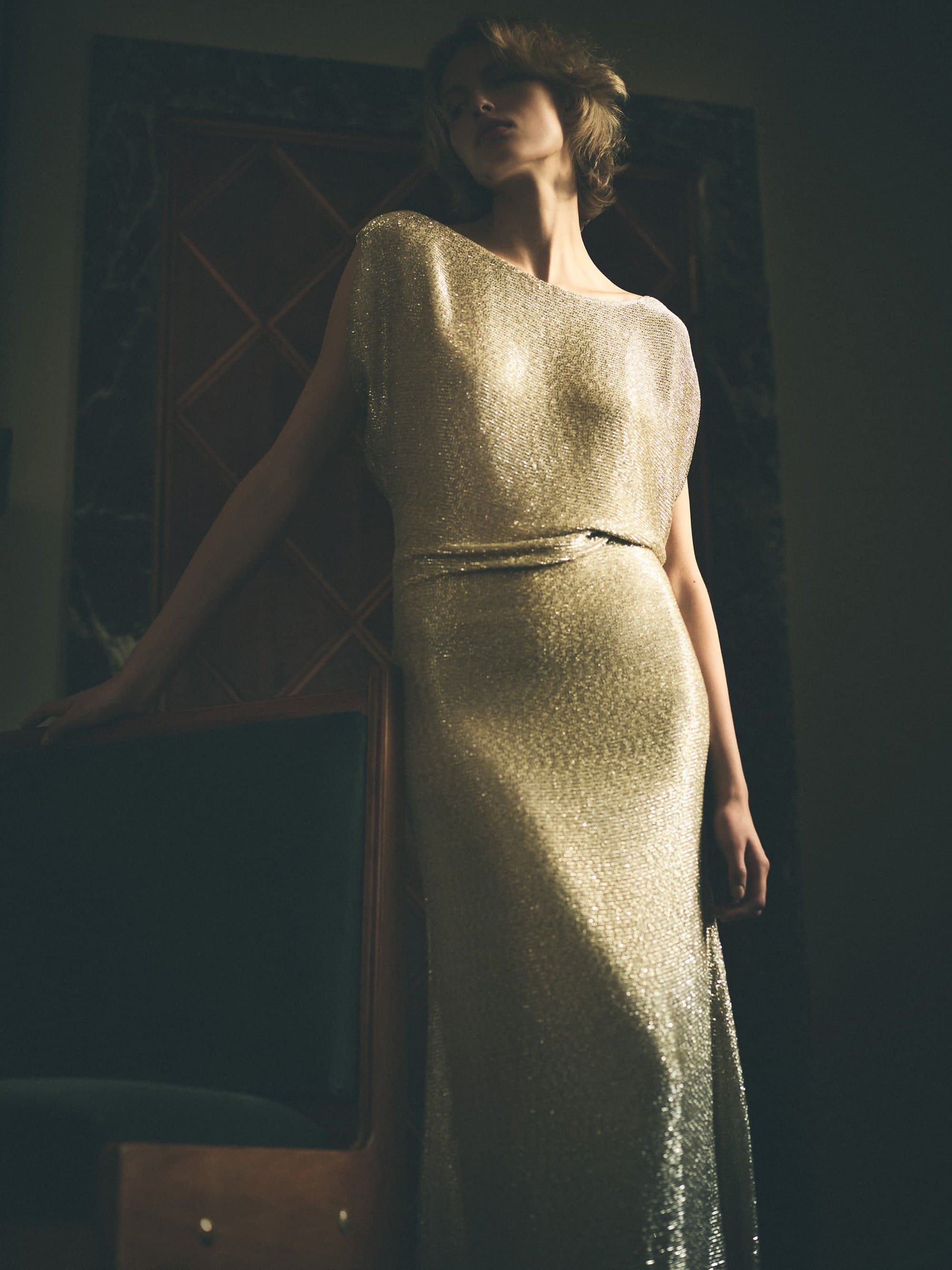 Model wearing a shimmering metallic gold evening gown posing indoors in a dimly lit room with dark wooden paneling.