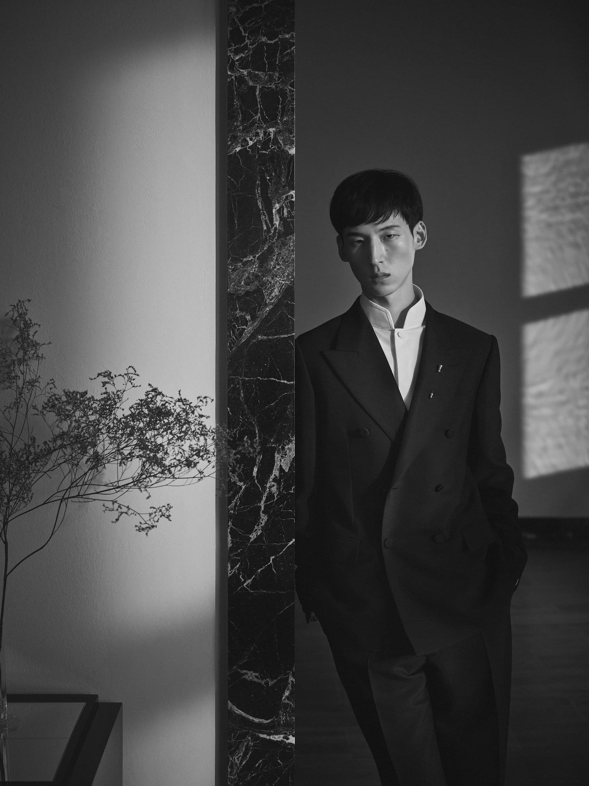 Man wearing a dark double-breasted suit leaning against a dark marble column in a high-contrast interior setting.