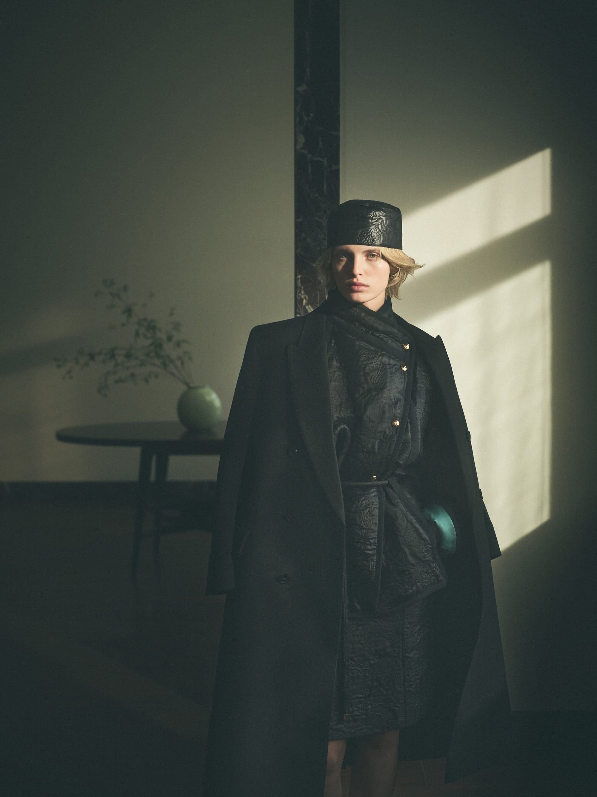 Model in dark coat and matching hat standing indoors near a table with a vase, illuminated by strong slanting light.