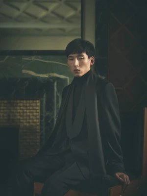 Man wearing a dark suit jacket and turtleneck seated indoors against a moody interior background.