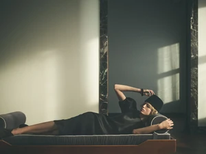 Model lying on a daybed wearing a dark dress and pillbox hat in a sunlit interior space