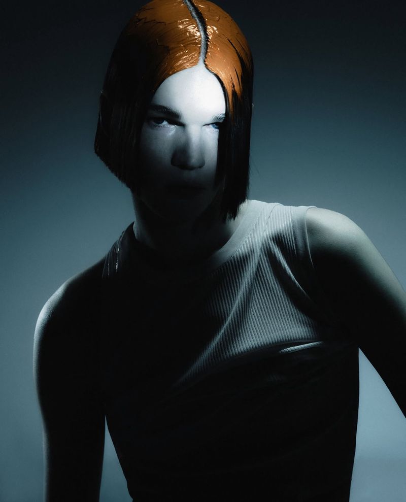 Stylish studio portrait of a person with dyed orange blunt bob haircut, wearing a ribbed tank top, against a graduated grey background.