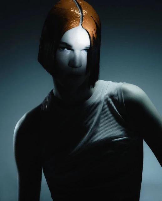 Stylish studio portrait of a person with dyed orange blunt bob haircut, wearing a ribbed tank top, against a graduated grey background.