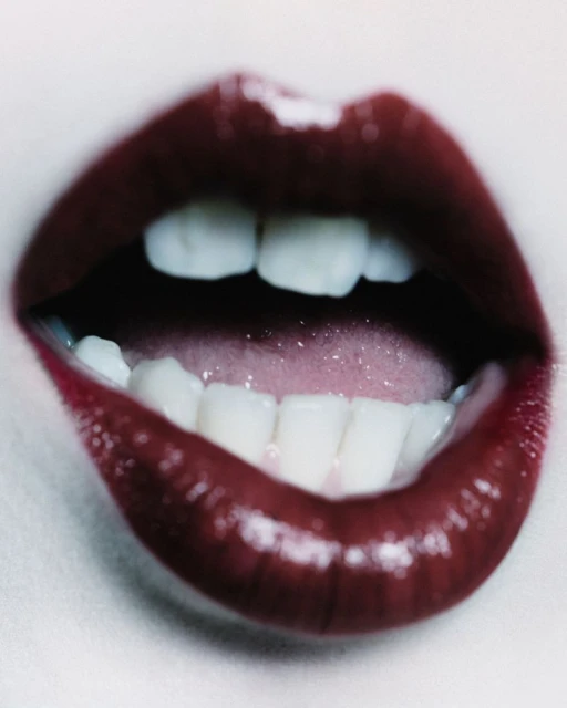 Close-up shot of a woman's open mouth with red lipstick, revealing white teeth and pink tongue, against a soft grey background.