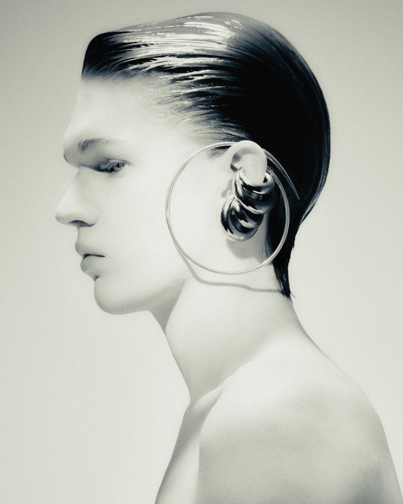 Monochrome portrait of a bare-chested adult male in profile, featuring slicked-back hair and large hoop earrings, showcasing beauty and fashion.