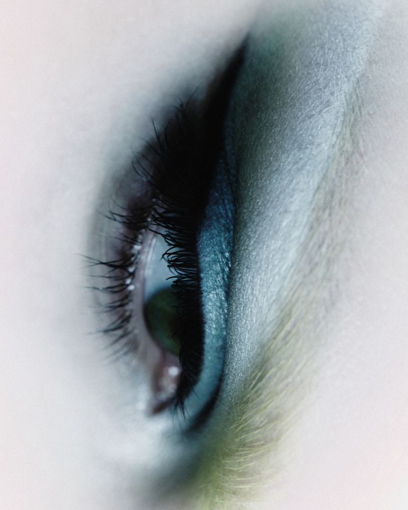 Macro shot of a single green eye with long black lashes and turquoise makeup, illuminated by soft, even lighting, emphasizing clarity and color.