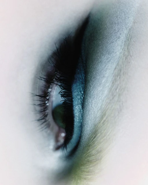 Macro shot of a single green eye with long black lashes and turquoise makeup, illuminated by soft, even lighting, emphasizing clarity and color.