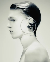 Profile portrait of a model with slicked-back wet-look hair wearing large metallic hoop earrings against a neutral light background.