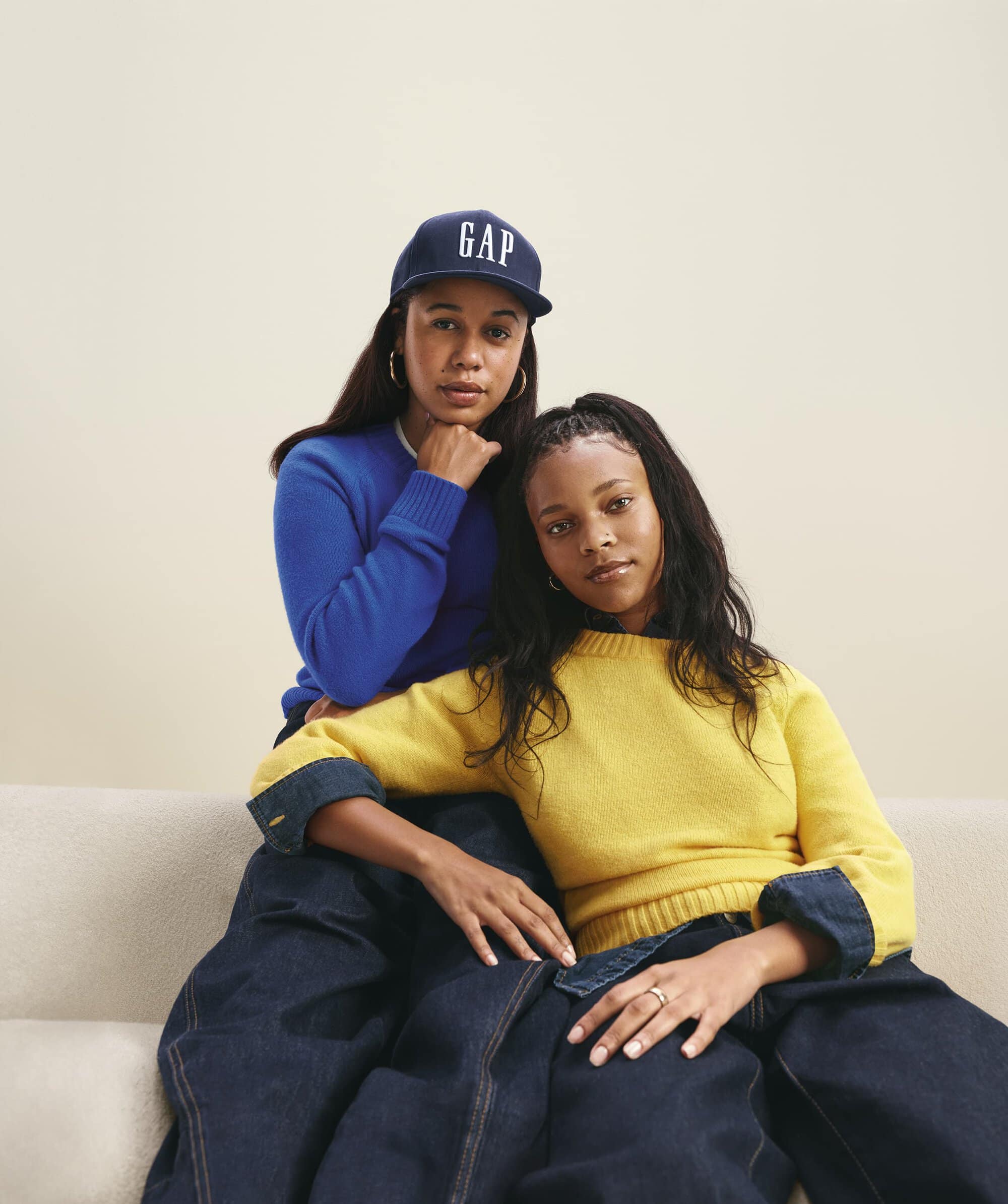 Two women seated together wearing knit sweaters and blue baseball cap against neutral studio background