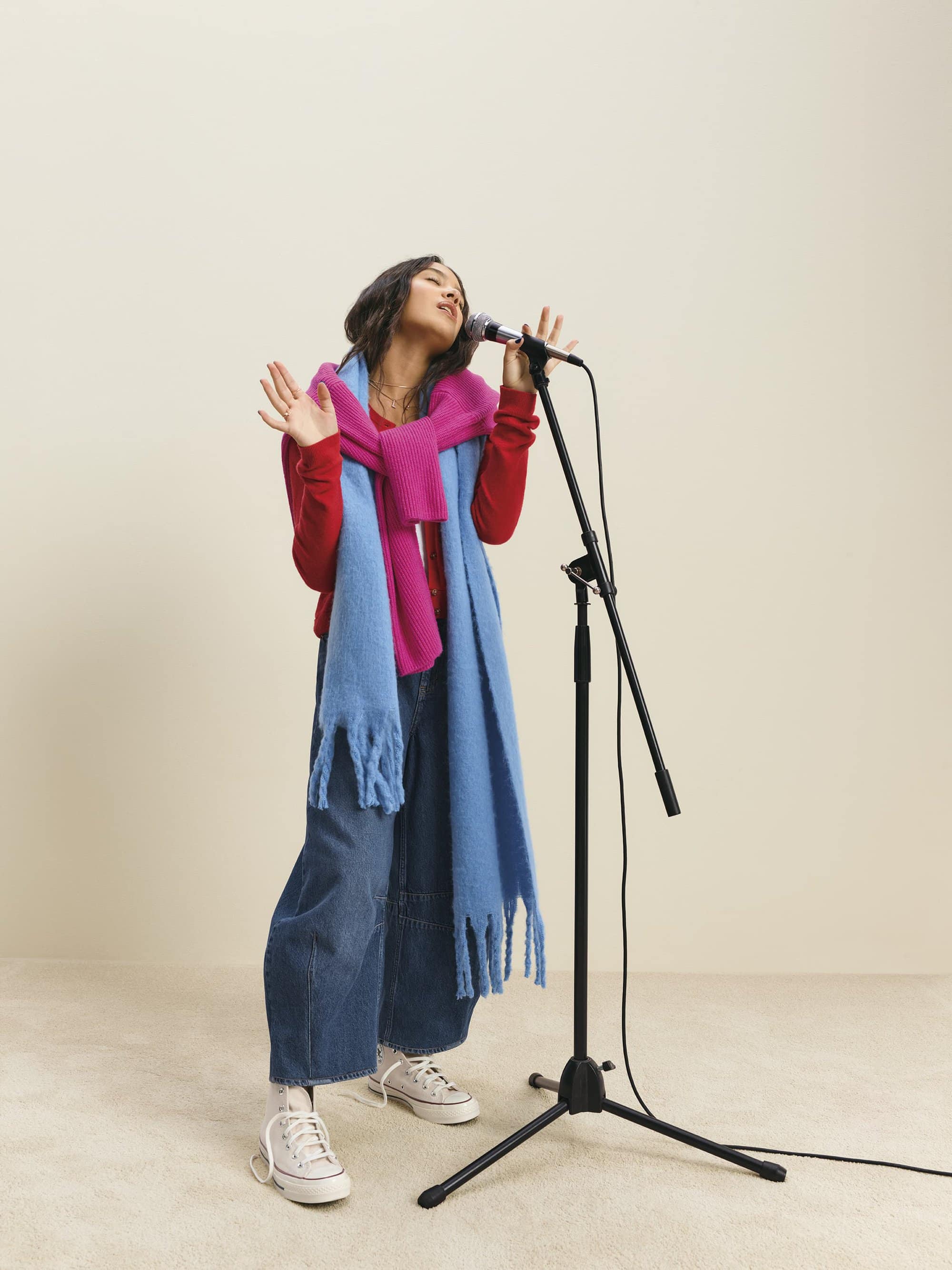 Woman singing into a microphone on a stand, wearing a red sweater, blue scarf, and denim jeans against a plain backdrop.
