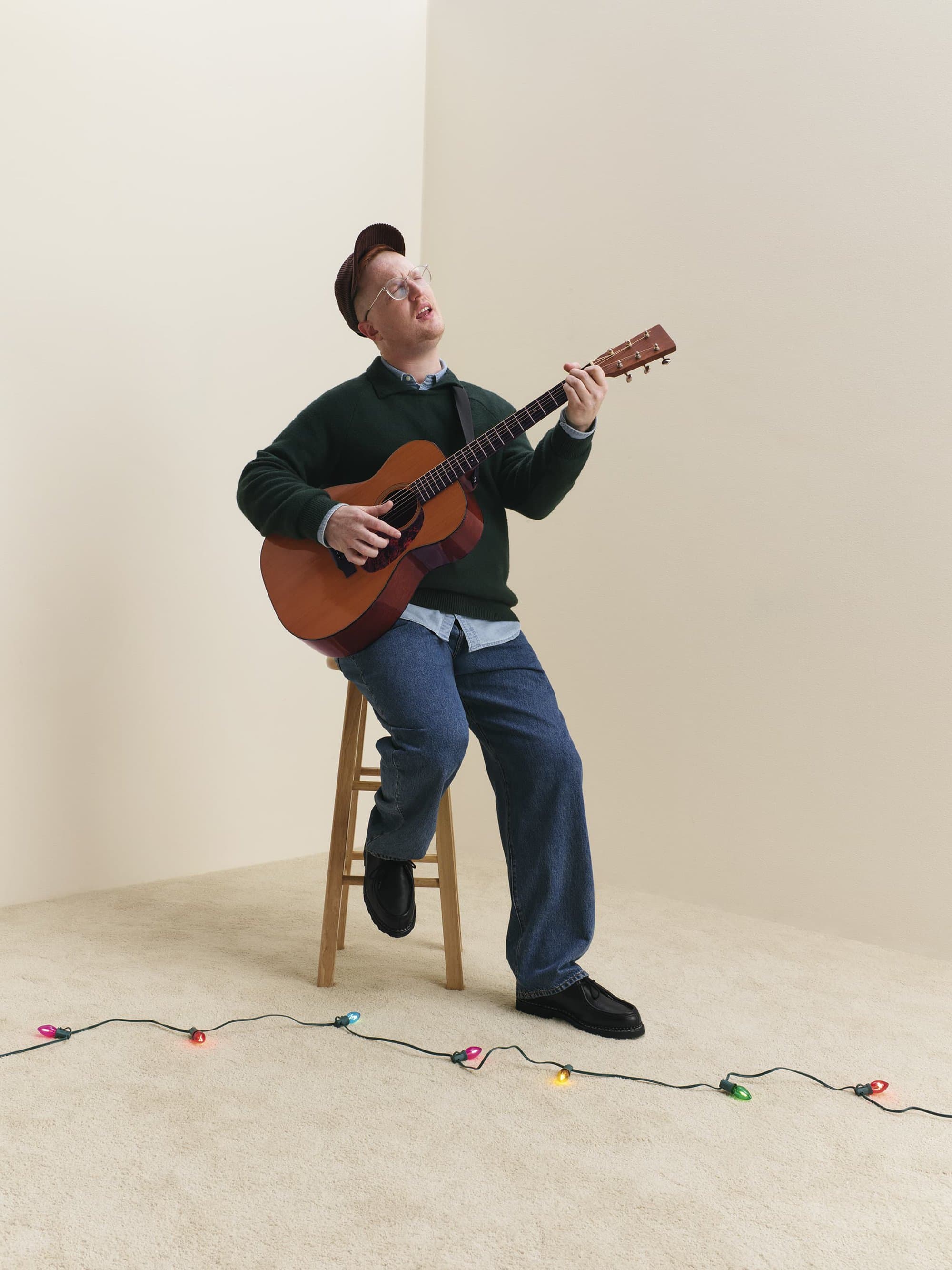 Man seated on wooden stool playing acoustic guitar against plain background with string lights