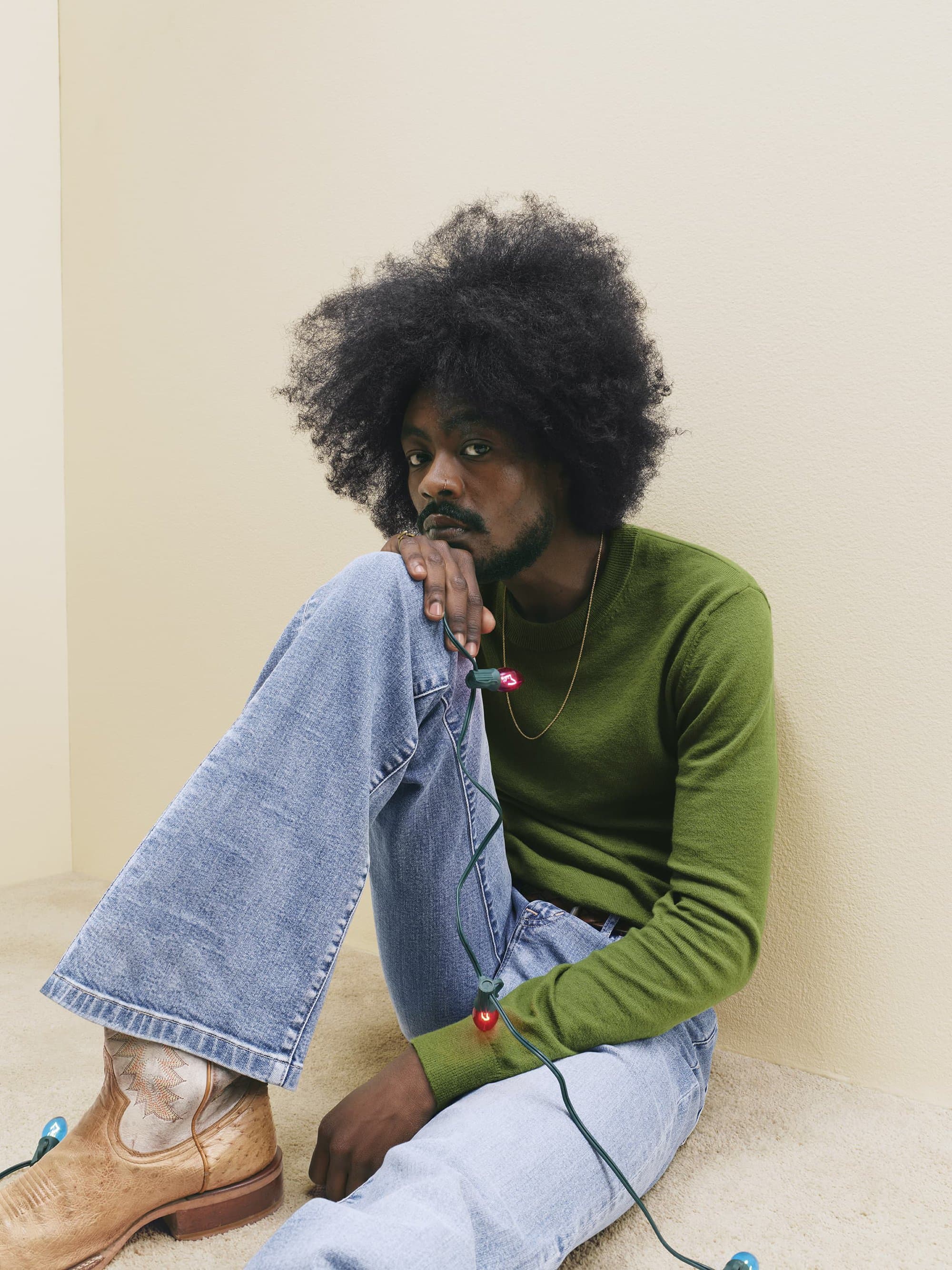 Black man with afro wearing a green sweater and light wash jeans seated against a pale wall holding holiday string lights.