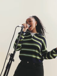 Black woman singing into a microphone wearing a striped polo sweater against a neutral background