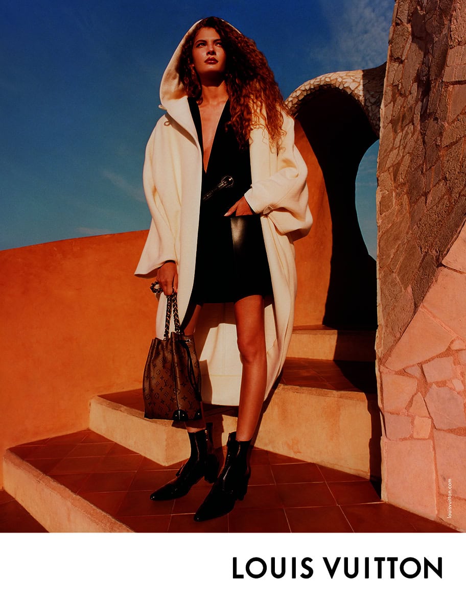 Model standing on stone steps wearing a hooded white coat and holding a bucket bag against a clear blue sky