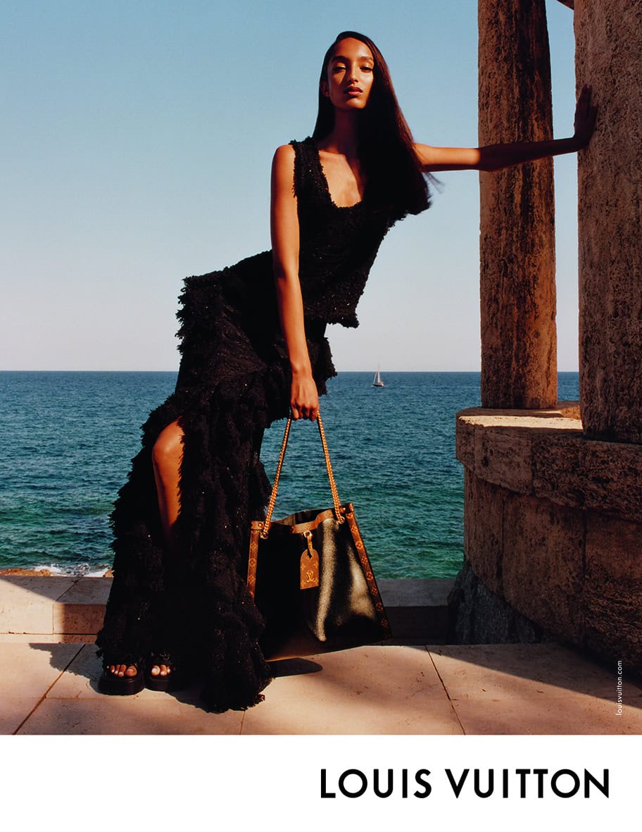 Model in black textured dress leaning against stone column holding monogram handbag with ocean background