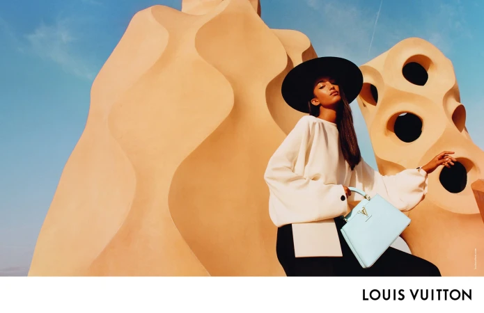 Model in wide-brimmed black hat and white top holding light blue handbag against sculptural architectural forms