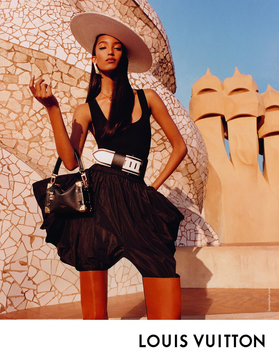 Model wearing a wide-brimmed hat, black top, and voluminous skirt, holding a handbag against distinctive mosaic architecture.