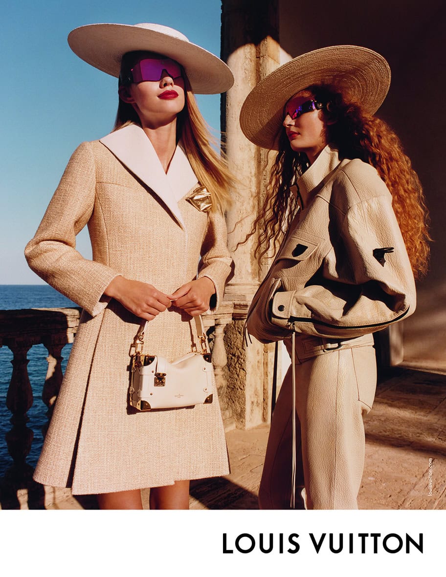 Two models wearing wide hats, sunglasses, and cream-colored luxury attire posing on a balcony overlooking the sea.
