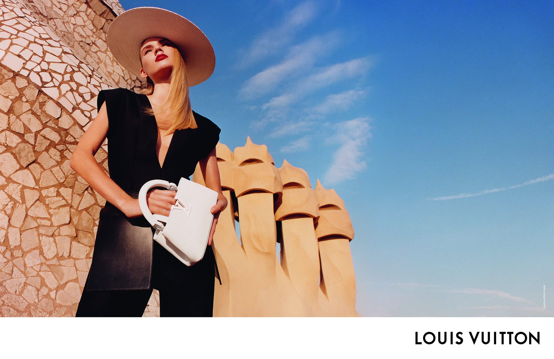 Model in black top and wide-brim hat holding a white handbag against a tiled wall and blue sky