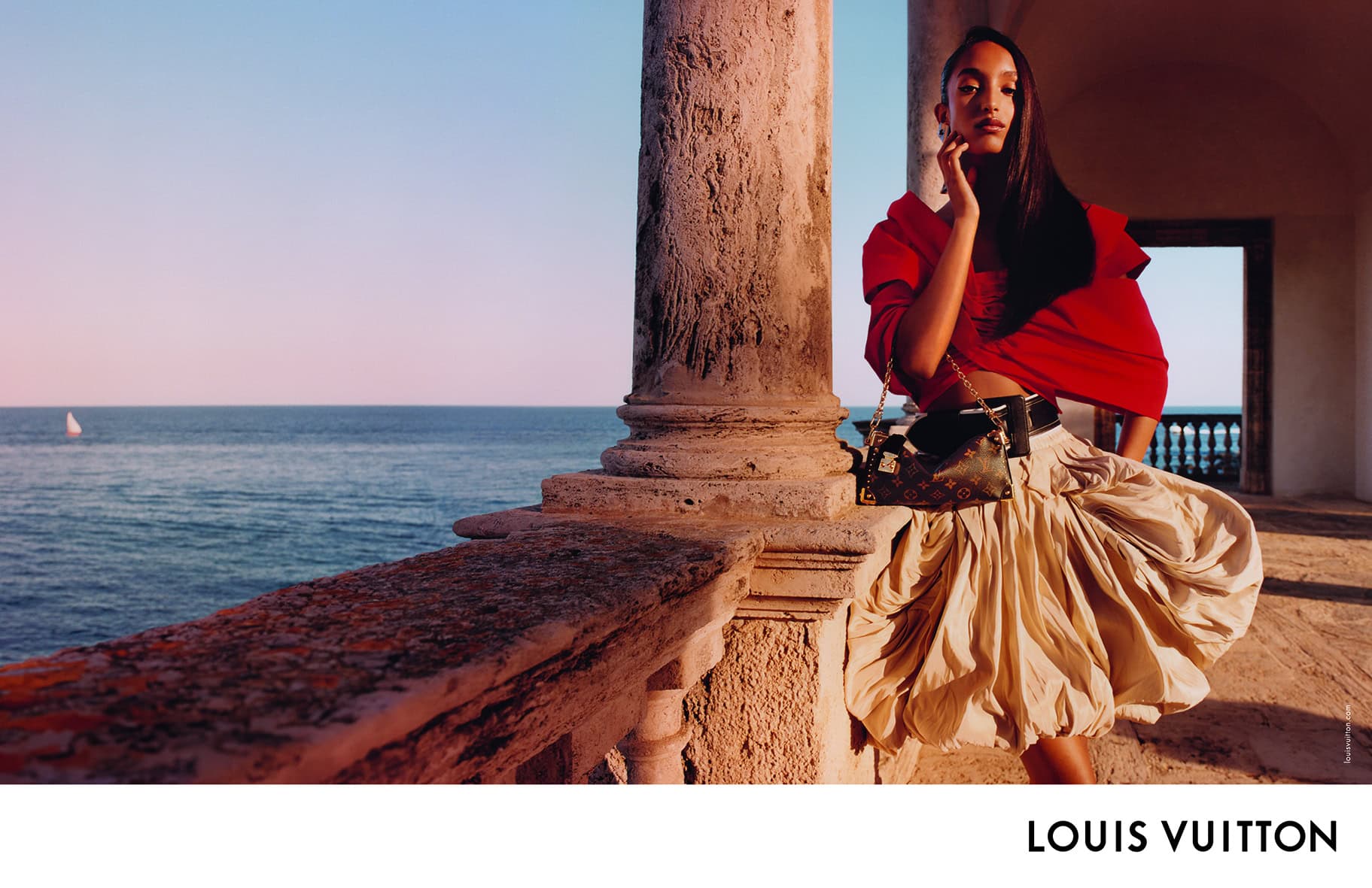 Model in red top and beige skirt holding a monogram handbag near a stone balcony overlooking the sea at sunset.