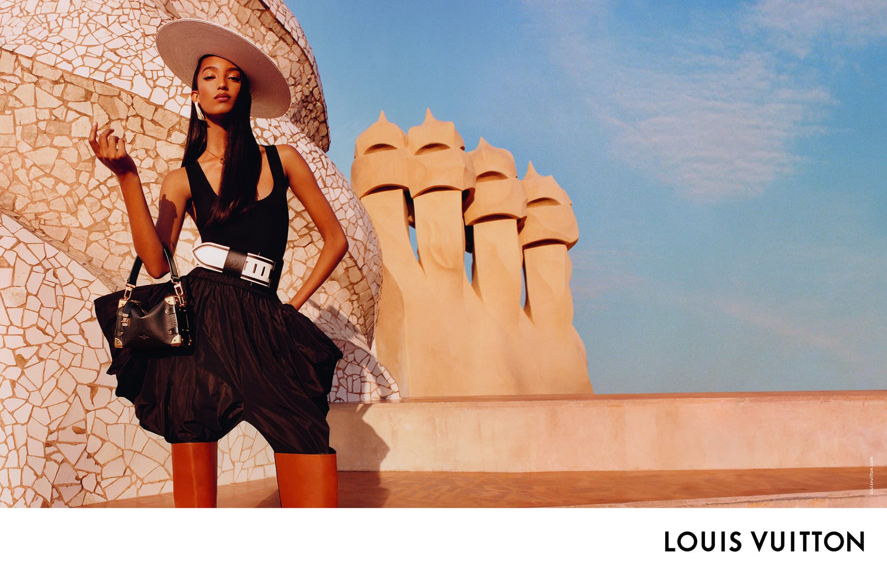 Model in a wide-brimmed hat, black top, and puffy trousers holding a handbag against an architectural rooftop background.