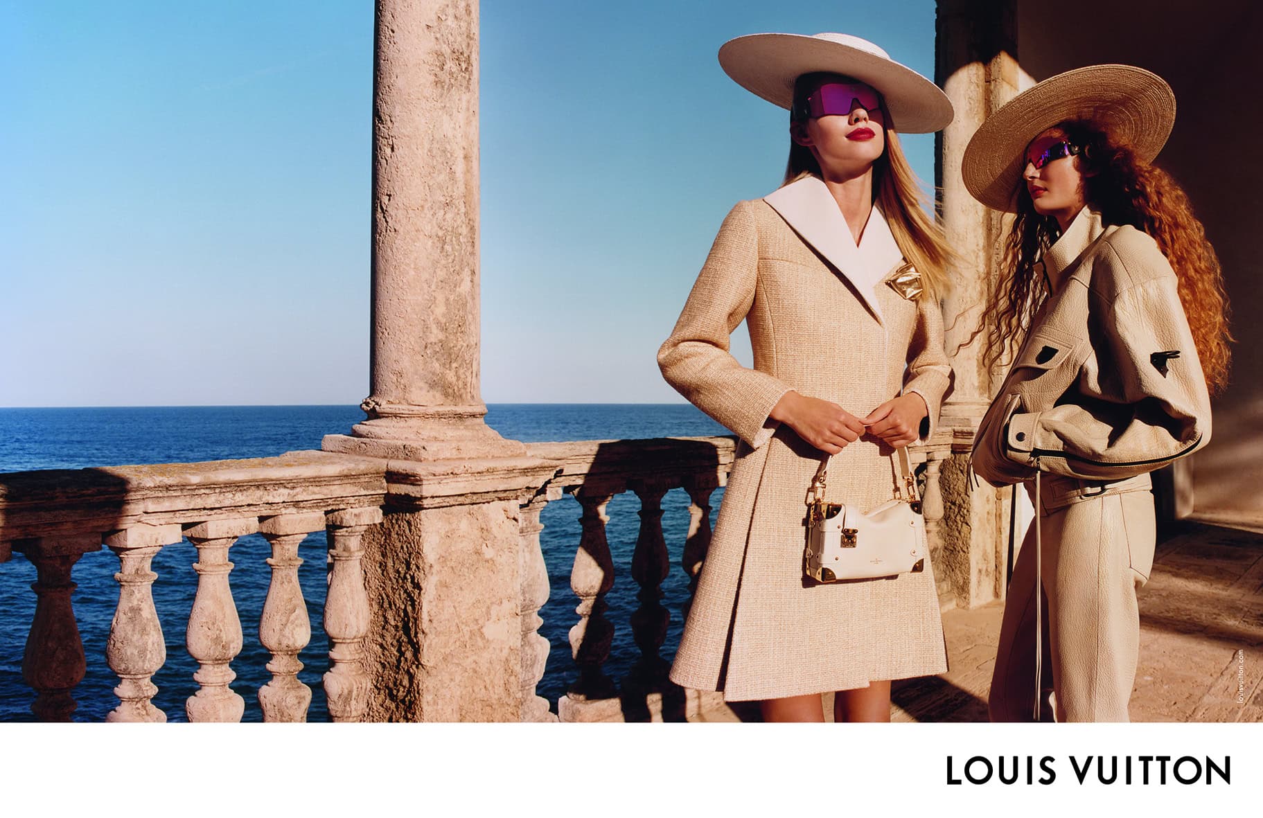 Two models wearing wide-brimmed hats and sunglasses standing on a stone terrace overlooking the blue sea