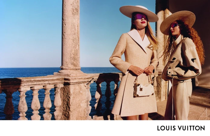 Two models wearing wide-brimmed hats and sunglasses standing on a stone terrace overlooking the blue sea