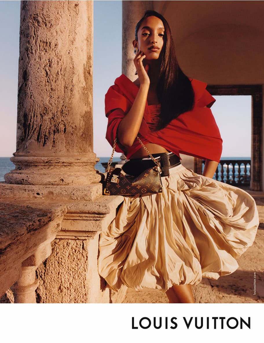 Model in red top and voluminous skirt holding a monogram handbag near stone columns with an ocean view for Louis Vuitton Cruise 2025.
