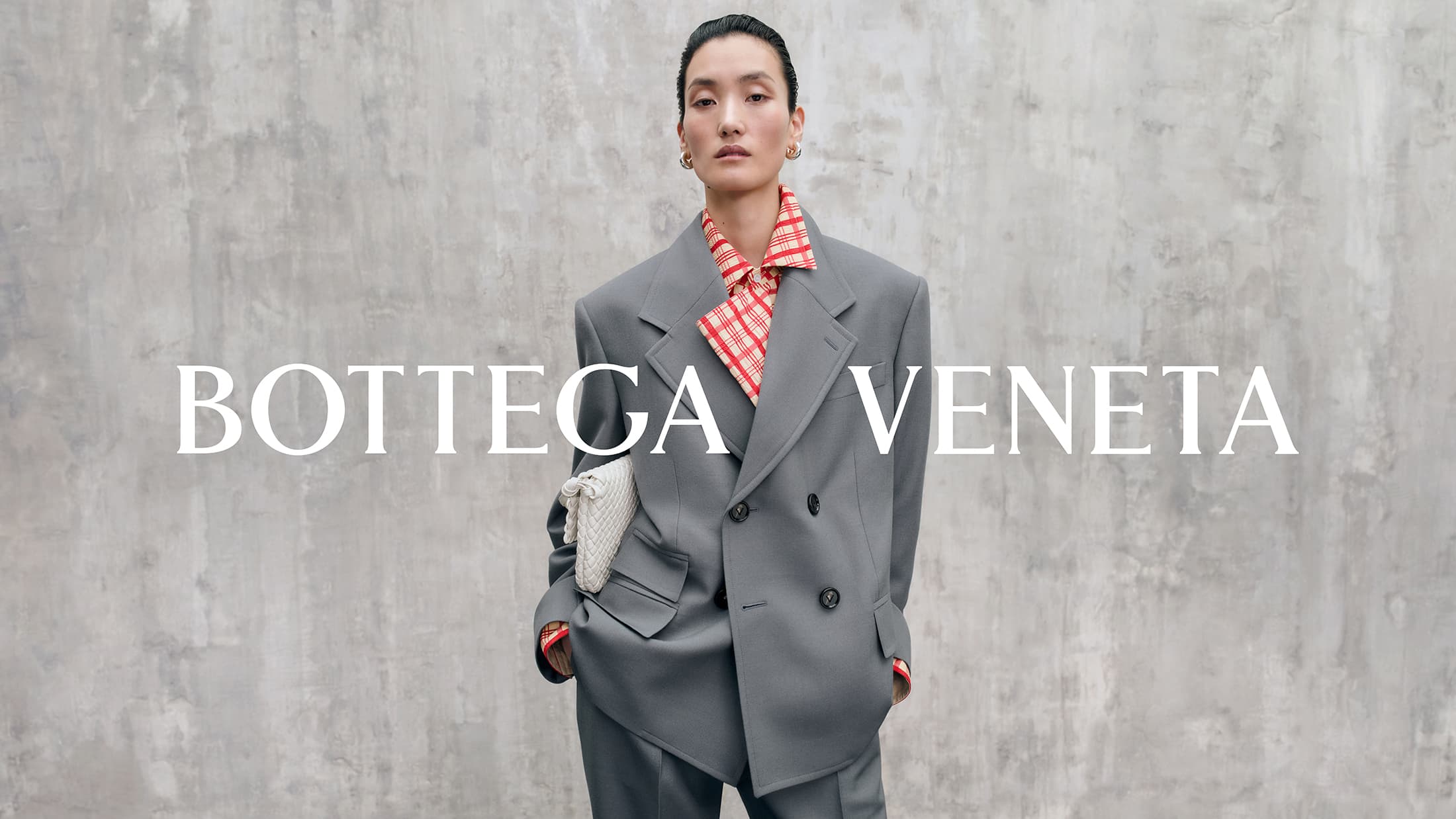 Model wearing an oversized grey double-breasted blazer and red patterned shirt against a textured concrete wall holding a white woven clutch