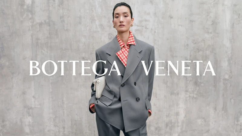 Model wearing an oversized grey double-breasted blazer and red patterned shirt against a textured concrete wall holding a white woven clutch