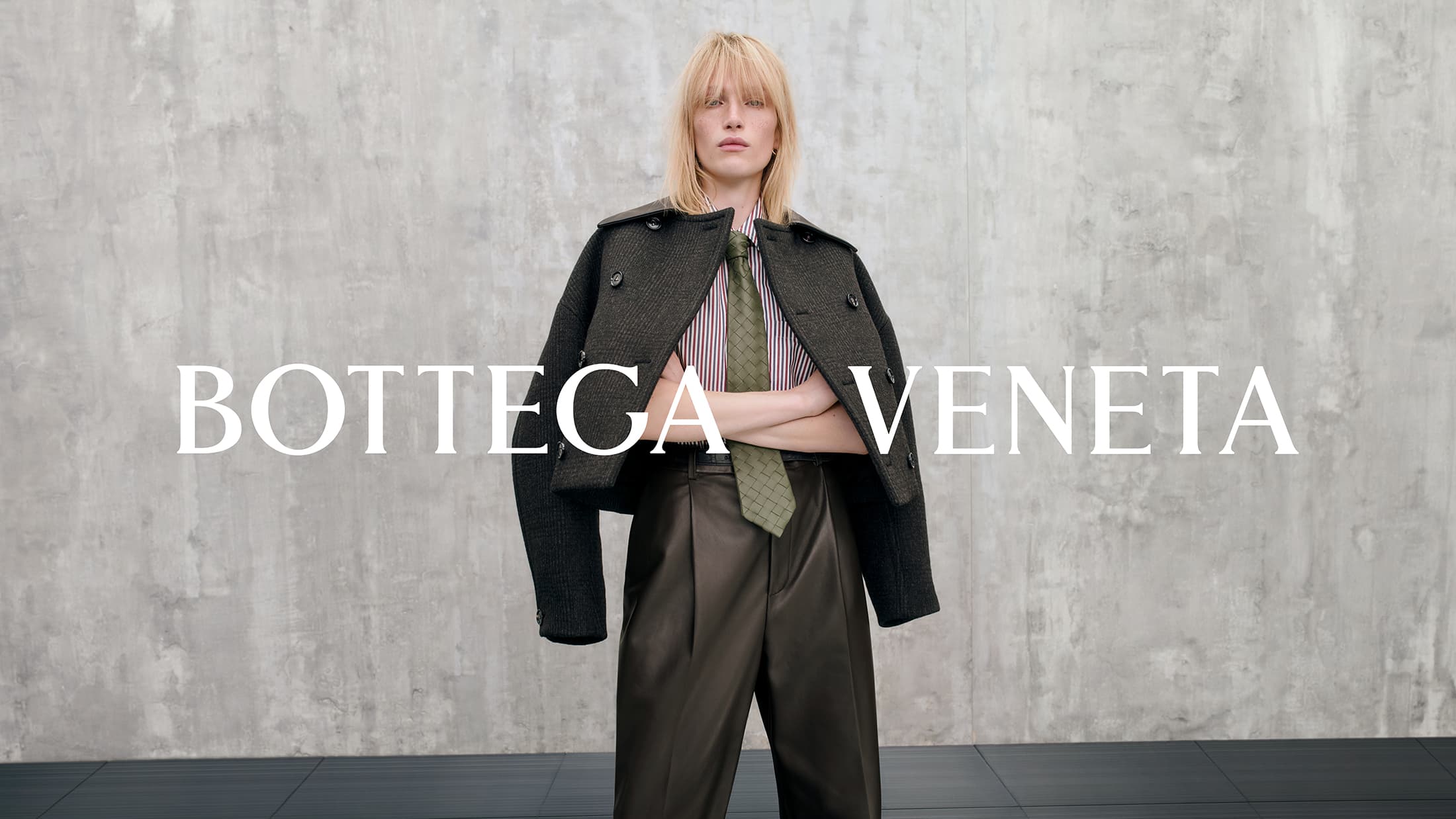 Model wearing a dark textured cropped coat, striped shirt, green tie, and dark trousers against a concrete wall background