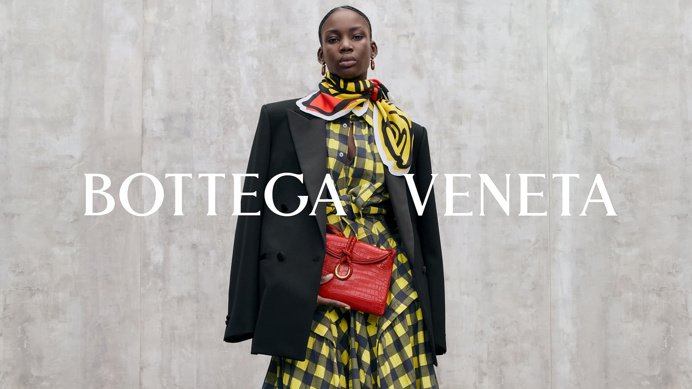 Model in black blazer and yellow plaid dress wearing a patterned scarf and holding a red handbag against a concrete wall