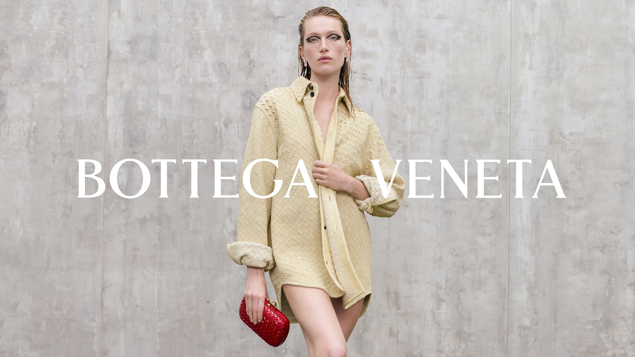 Model in a pale woven shirt holding a glossy red clutch against a concrete wall for Bottega Veneta Pre-Spring 2025 campaign