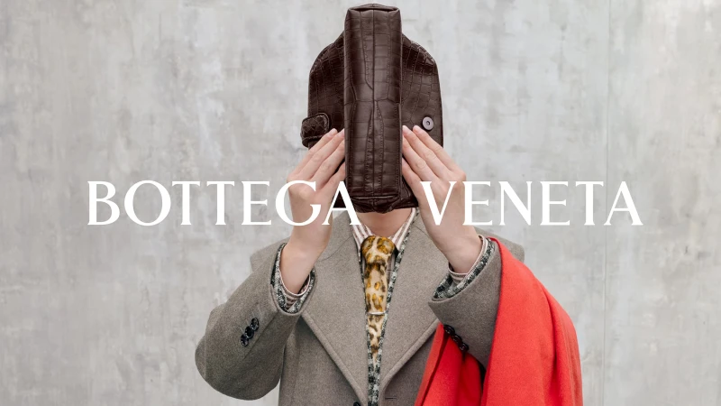 Model holding a dark brown crocodile leather handbag over their face, wearing a grey coat, leopard tie, and holding a red coat.