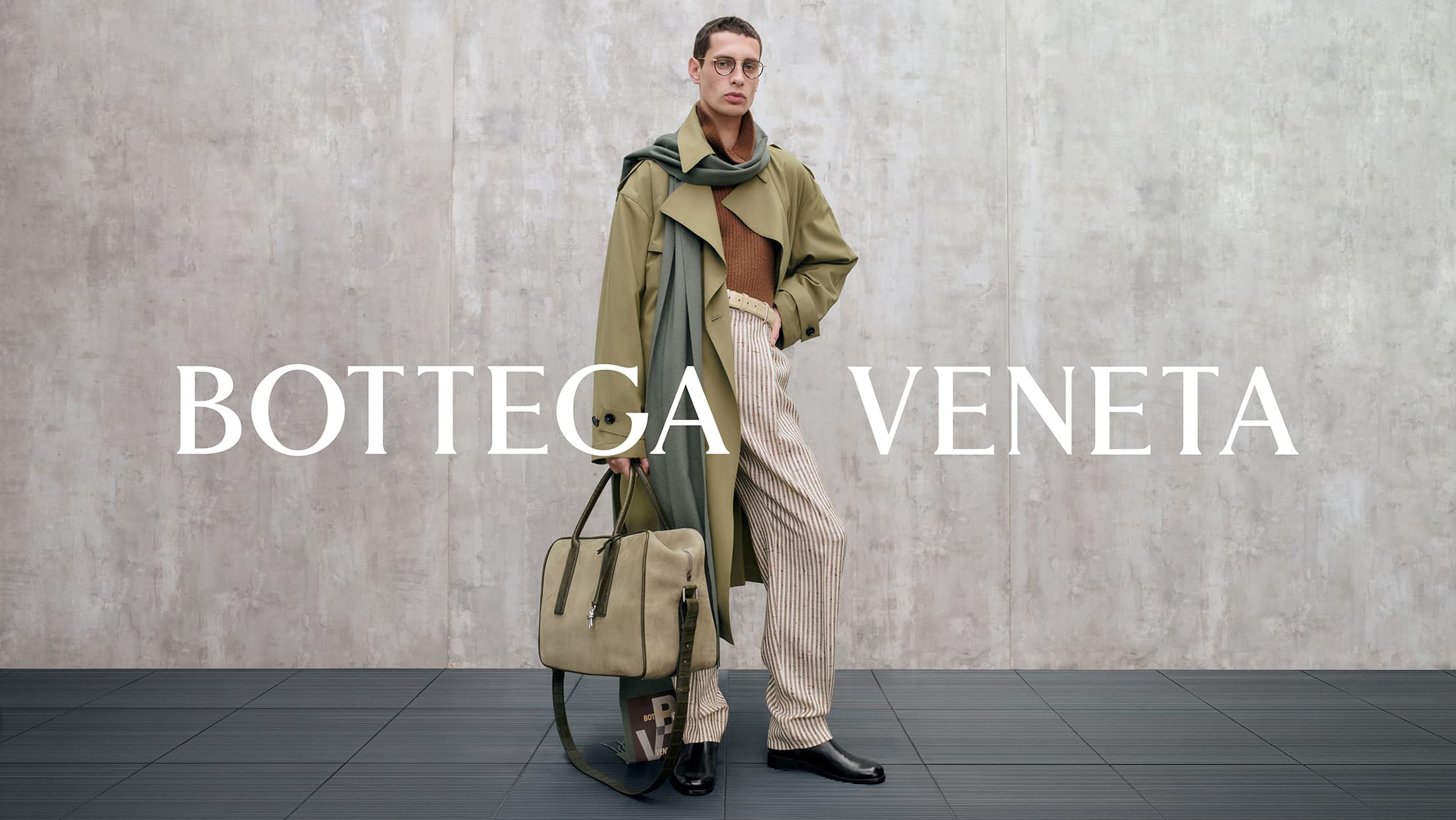 Model wearing an olive coat, striped trousers, and holding a large bag against a textured grey wall