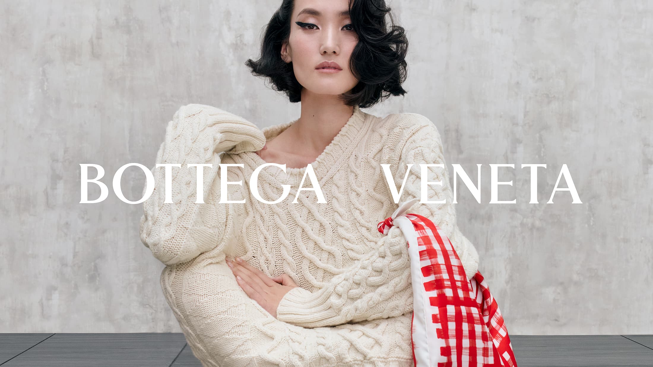Model in a cream cable-knit sweater holding a red and white checkered scarf against a concrete wall