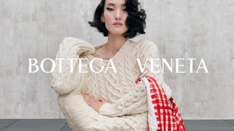 Model in a cream cable-knit sweater holding a red and white checkered scarf against a concrete wall