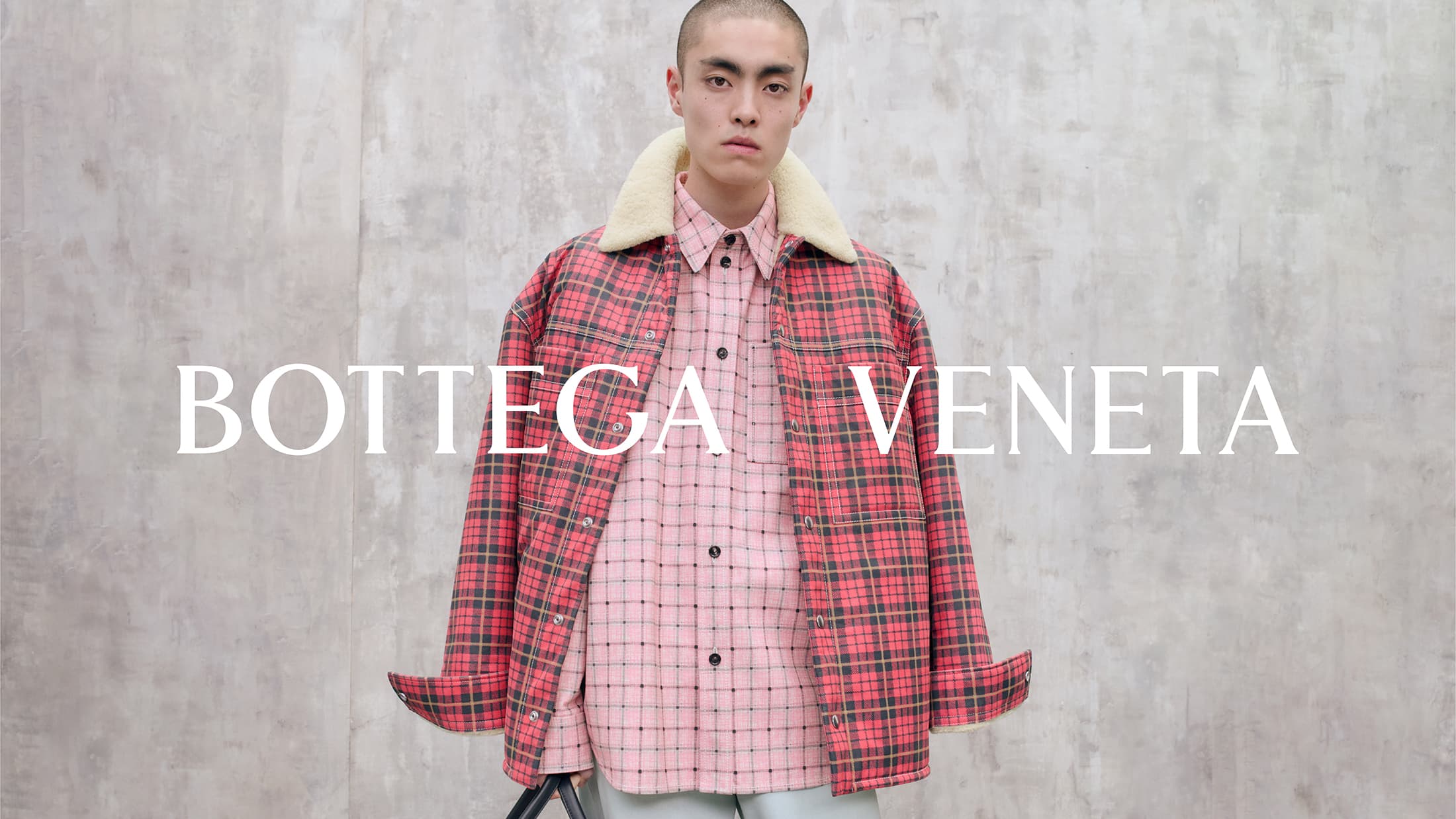 Model wearing a plaid jacket with a shearling collar and pink patterned shirt against a textured grey wall