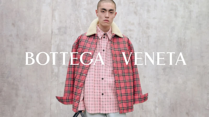 Model wearing a plaid jacket with a shearling collar and pink patterned shirt against a textured grey wall