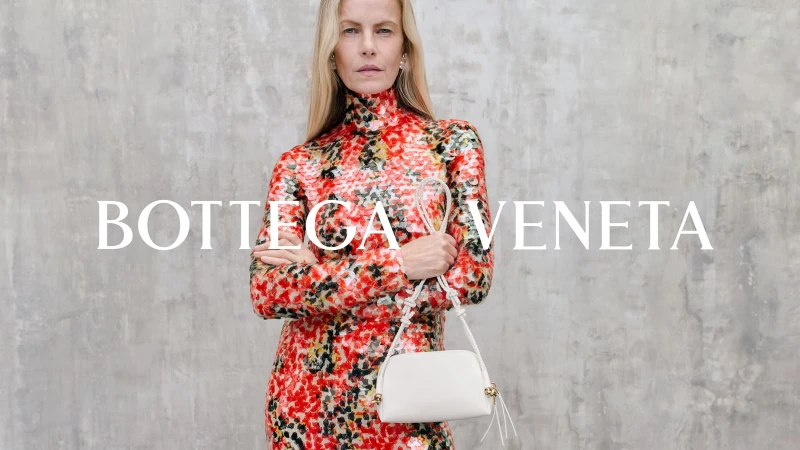 Woman in a red patterned turtleneck dress holding a white shoulder bag against a textured grey wall