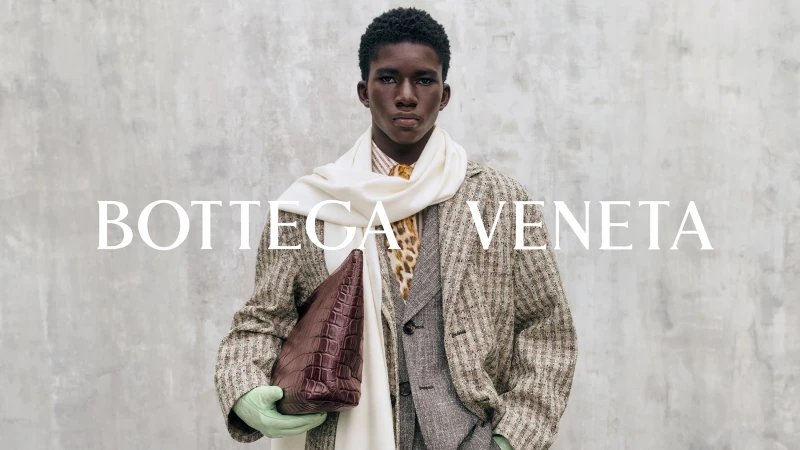 Black man wearing a textured grey coat, white scarf, and holding a brown leather handbag against a grey concrete wall