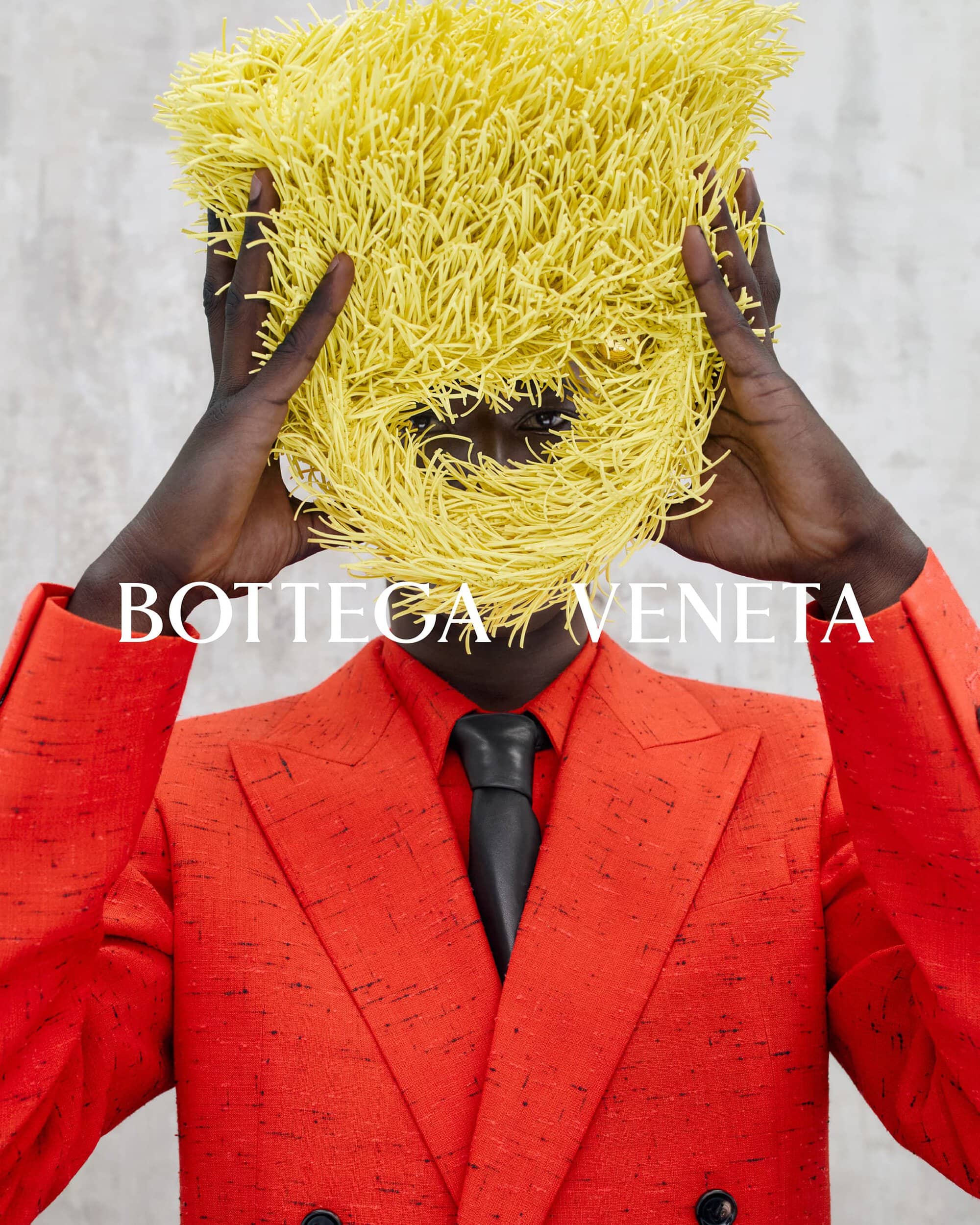 Model holding a textured yellow fringed headpiece, wearing an orange blazer and black tie against a white seamless background