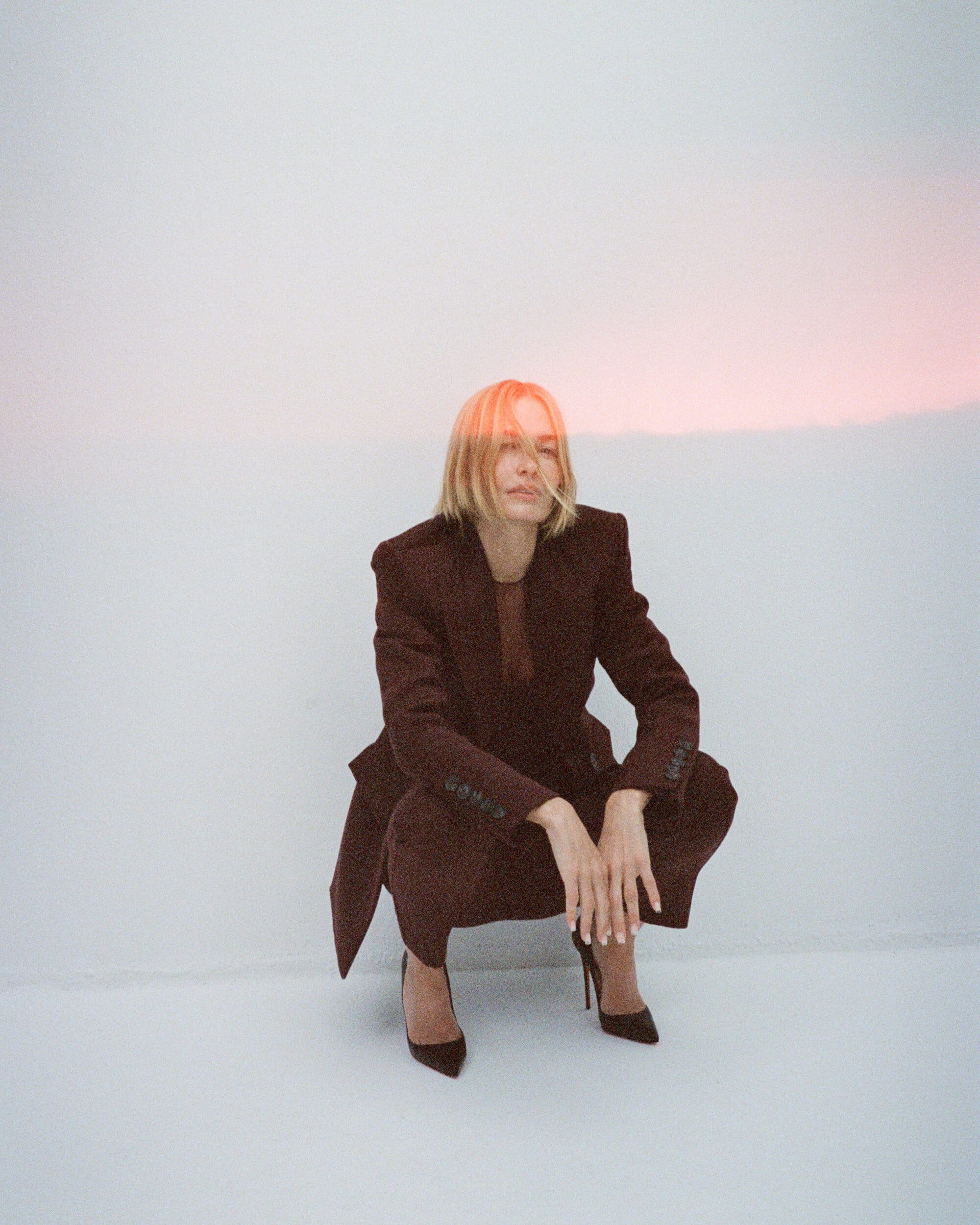 Model in burgundy tailored pantsuit crouching against a pale wall with soft pink lighting across her head