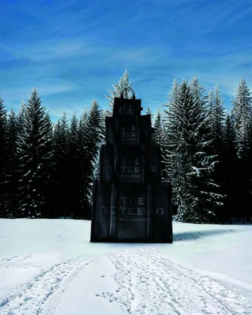 Giant stack of black leather tote bags standing upright in a snowy winter landscape surrounded by pine trees under a clear blue sky