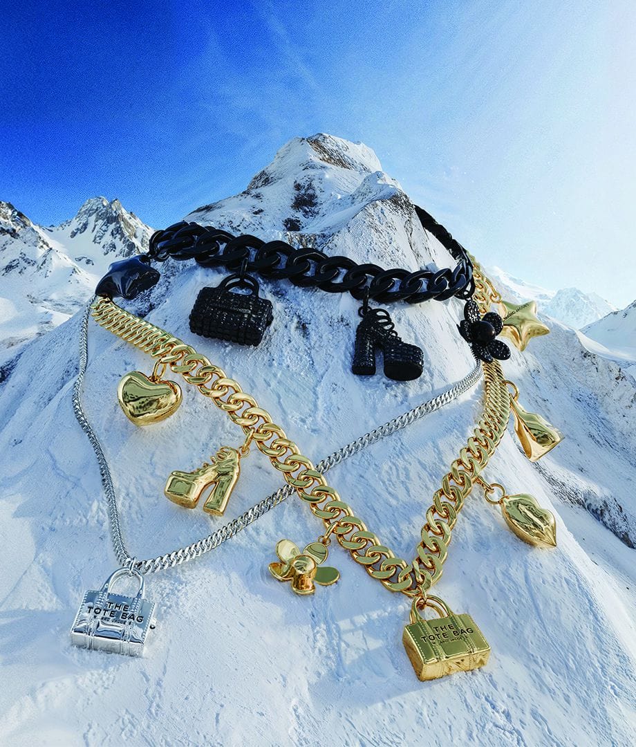 Chunky gold, silver, and black charm necklaces draped over a snow-covered mountain peak under a clear blue sky.