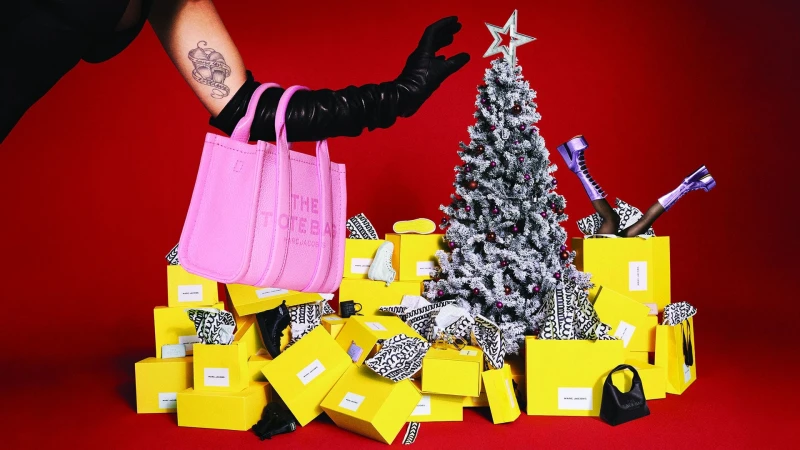 Pink Marc Jacobs tote bag held by a gloved hand over a pile of yellow gift boxes and a frosted Christmas tree against red