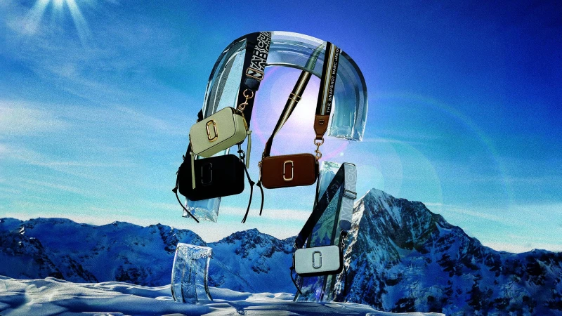 Marc Jacobs handbags suspended on clear acrylic arches against a snowy mountain landscape under a bright blue sky.