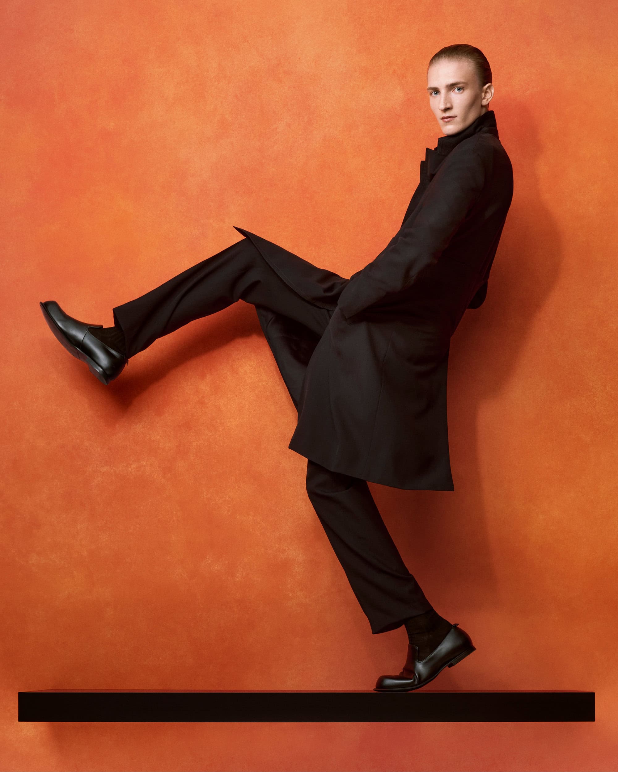 Man wearing a black coat and trousers kicking his leg up against an orange textured wall
